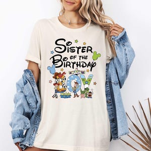 A woman wearing a white t-shirt with a graphic design featuring characters from the Toy Story movie, along with the text "Sister of the Birthday Joy".