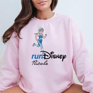 A woman wearing a pink sweatshirt with a Disney-themed graphic and text.