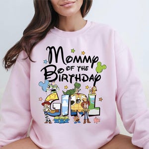A woman wearing a pink sweatshirt with a cartoon character design and text that says "Mommy of the Birthday Girl".