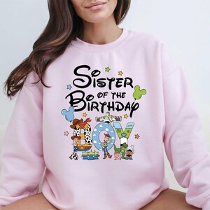 A woman wearing a pink sweatshirt with a cartoon character design and text that says "Sister of the Birthday".