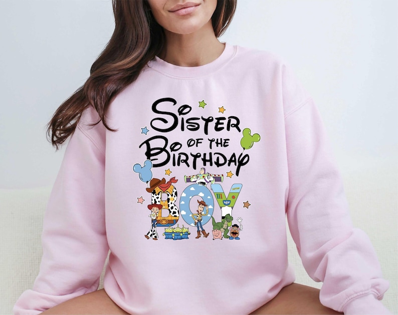 A woman wearing a pink sweatshirt with a cartoon character design and text that says "Sister of the Birthday".