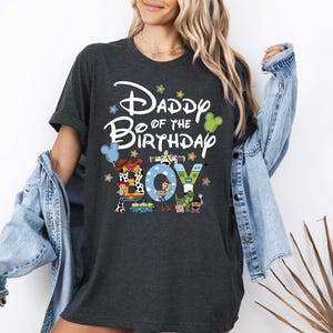 A woman wearing a black t-shirt with a Disney-themed design and text, standing in front of a white wall.
