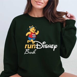 A woman wearing a green sweatshirt with a Winnie the Pooh character and the text "runDisney Bob" printed on it.