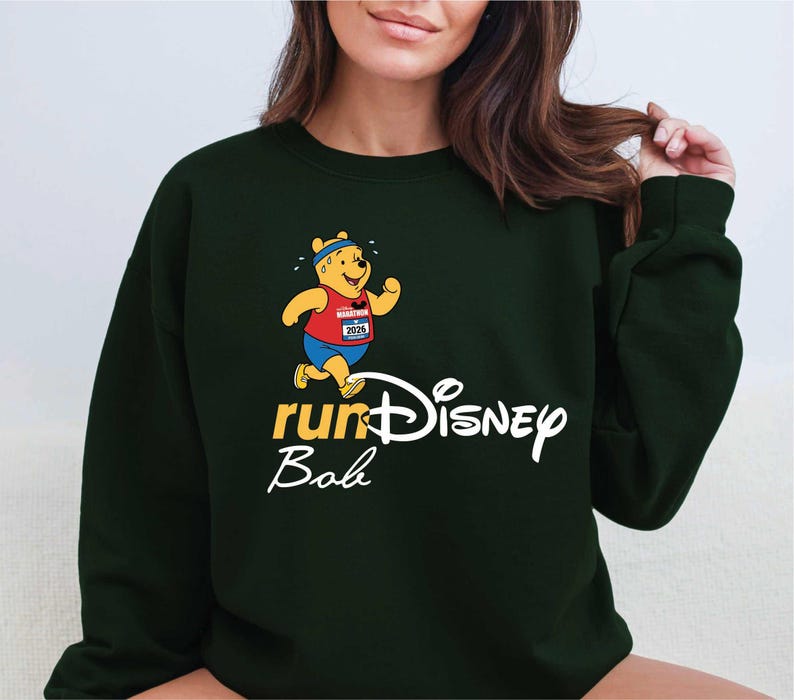 A woman wearing a green sweatshirt with a Winnie the Pooh character and the text "runDisney Bob" printed on it.