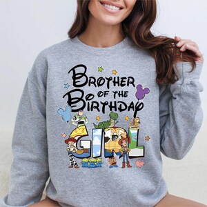 A woman wearing a gray sweatshirt with a colorful graphic design that says "Brother of the Birthday Girl" featuring characters from the Toy Story movie.