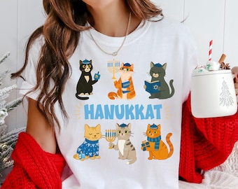 May include: White t-shirt featuring six cartoon cats celebrating Hanukkah. Each cat wears a yarmulke and holds Hanukkah symbols like a menorah and dreidel. The word "HANUKKAT" is printed in blue below the cats.