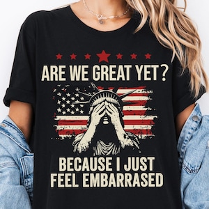 May include: Black t-shirt with a graphic of the Statue of Liberty covering her eyes with her hands. The graphic is in front of an American flag with the text "Are we great yet? Because I just feel embarrassed".