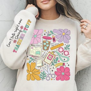 May include: A beige sweatshirt with a graphic print of colorful teacher-themed items, including coffee cups, books, pencils, and flowers. The text "Can't talk right now *Doing Teacher Stuff*" is printed on the sleeve.