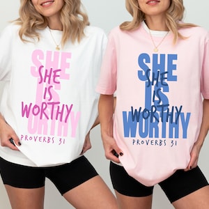 May include: Two women wearing t-shirts with the text "SHE IS WORTHY" in pink and blue. The white shirt has pink text, and the pink shirt has blue text. The text "PROVERBS 31" is below the main text. Both women are wearing black shorts.