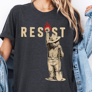Op de afbeelding: Een donker grijs t-shirt met een afbeelding van een beer die een rangerhoed draagt en een schop vasthoudt. De beer steekt zijn vuist in de lucht met vlammen die uit zijn hand komen. Het woord "RESIST" staat boven de beer gedrukt.