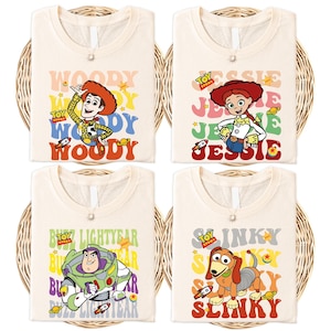 May include: Four cream-coloured t-shirts featuring characters from the Toy Story film. Each shirt displays a different character: Woody, Jessie, Buzz Lightyear, and Slinky Dog, with their names in colourful, retro-style text. The shirts are folded in a woven basket.