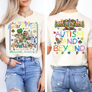 Peut inclure: T-shirt crème avec texte et graphiques colorés promouvant la sensibilisation à l'autisme. Le devant présente des personnages de dessins animés et les mots "Autisme Conscience et au-delà". Le dos affiche "Autisme et au-delà" avec une fusée et des pièces de puzzle.