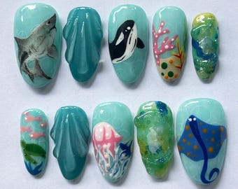 Puede incluir: Un conjunto de diez uñas artificiales con temática oceánica. Los diseños incluyen un tiburón, una orca, coral, una medusa y una manta raya, todo sobre un fondo azul claro. Las uñas varían en forma y tamaño, algunas con elementos 3D.