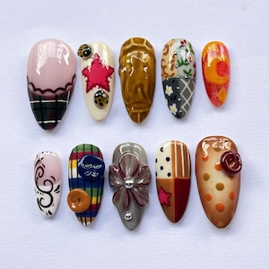 May include: Ten artificial nails with diverse designs. The nails feature patterns such as plaid and floral, alongside embellishments like buttons and a bow. The colour palette includes pink, green, brown, and orange, with varied textures.