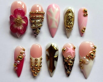 May include: Ten pink and white false nails with various gold and pearl embellishments. The nails feature designs including a flower, a cross, a star, and a religious figure.