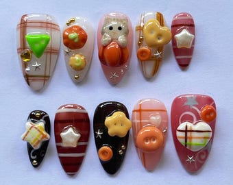 May include: A set of ten artificial nails with various designs. The nails feature fall-themed decorations, including pumpkins, stars, and a heart. Colors include orange, brown, pink, and white. Some nails have a plaid pattern, and others have 3D embellishments.