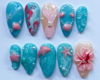 Puede incluir: Un conjunto de diez uñas artificiales con temática veraniega. Las uñas presentan una variedad de diseños, incluyendo conchas marinas, estrellas de mar, flores de hibisco y efectos de gotas de agua. Los colores incluyen tonos de azul, rosa y blanco.