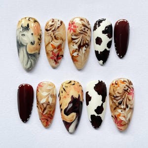 May include: A collection of ten artificial nails with various designs. The designs include a horse portrait, floral patterns, and a cow print. The nails are painted in shades of brown, beige, white, and burgundy, with some featuring gold accents.