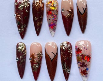 May include: A set of ten artificial nails in various designs. The nails feature a burgundy base with gold leaf accents, glitter, and rhinestones. Some nails have a nude base with brown and black designs. The nails are almond-shaped.