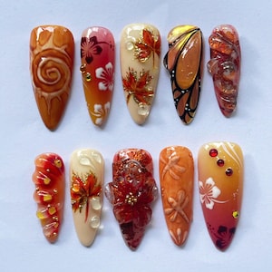 Puede incluir: Un conjunto de diez uñas artificiales con varios diseños temáticos de otoño. Las uñas presentan colores como naranja, rojo y dorado, con obras de arte detalladas que incluyen hojas, flores y alas de mariposa. Algunas uñas tienen adornos 3D.