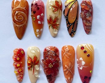 May include: A set of ten artificial nails with various autumn-themed designs. The nails feature colors like orange, red, and gold, with detailed artwork including leaves, flowers, and butterfly wings. Some nails have 3D embellishments.