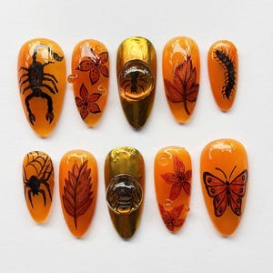 Könnte beinhalten: Ein Set aus zehn mandelförmigen Press-on-Nägeln in leuchtendem Orange. Die Designs zeigen schwarze Illustrationen von Skorpion, Spinnennetz, Tausendfüßler, Schmetterling und Blättern sowie rote Blumenmuster und zwei goldene Nägel mit 3D-Insekten-Charms.