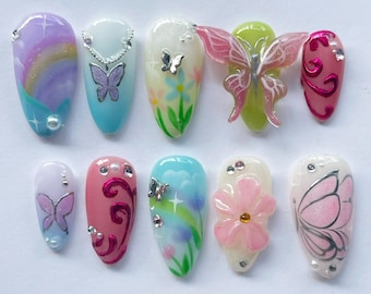 May include: A set of ten artificial nails with various designs. The nails feature pastel colors, butterflies, flowers, and decorative gems. Some nails have 3D butterfly embellishments, while others showcase painted floral and butterfly patterns.