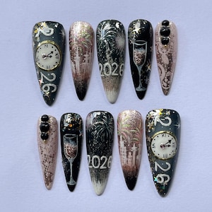 May include: A set of ten long, almond-shaped artificial nails with a New Year's Eve theme. Designs include clocks, fireworks, champagne glasses, and the year 2026. Colors include black, gold, silver, and various shades of pink and brown.