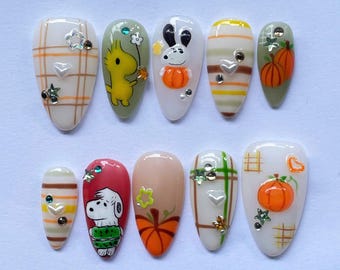May include: A set of ten almond-shaped false nails with autumn-themed designs. The nails feature cartoon characters, pumpkins, plaid, and stripes in colors like orange, green, and beige. Some nails are adorned with small, sparkly embellishments.