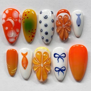 May include: A collection of ten artificial nails with various designs. The nails feature bright colors and patterns, including orange, yellow, white, and blue. Designs include fruit slices, fish, polka dots, and bow shapes. The nails are oval-shaped.