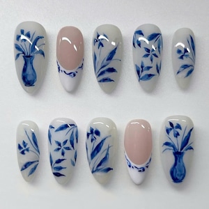 Puede incluir: Un conjunto de diez uñas postizas ovaladas. Las uñas están pintadas con una base blanca y presentan diseños florales azules, incluyendo jarrones y patrones botánicos. Dos uñas tienen un centro rosa con un borde blanco y azul.