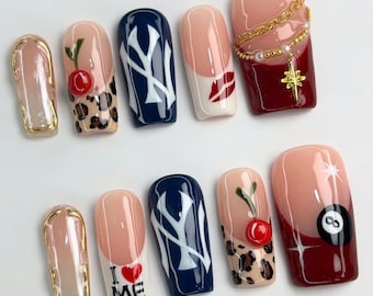 May include: Ten false fingernails with different designs.  The designs include a cherry, a leopard print, a New York Yankees logo, a red lip, a gold chain with a star, a 8-ball, and the words "I love me".