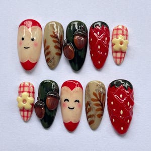 Könnte beinhalten: Ein Set aus zehn künstlichen Nägeln mit verschiedenen Designs. Die Designs umfassen Erdbeeren, Eicheln und lächelnde Gesichter. Einige Nägel weisen ein rot-weißes Vichy-Muster mit einer gelben Blume auf. Die Nägel sind in Rot-, Grün-, Beige- und Weißtönen lackiert.