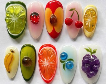 May include: A collection of ten artificial fingernails, each uniquely decorated with 3D fruit designs. The designs include a lime, strawberry, peach, cherries, lemon, banana, avocado, orange slice, blueberries, and grapes. The nails are painted in various colors.