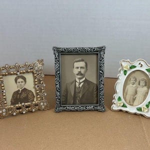 May include: Three decorative picture frames: a square gold-tone frame with embedded clear crystals, a rectangular dark silver-tone frame with an ornate filigree border, and a white ceramic oval frame featuring small sculpted floral accents.