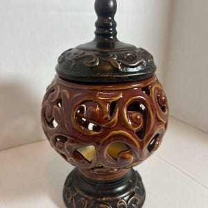 May include: A decorative ceramic jar with a lid. The jar is a deep reddish-brown color with intricate cut-out patterns and a dark brown base and lid. The lid has a small, rounded knob.