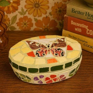 May include: An oval-shaped decorative box featuring a mosaic tile design. The lid displays a central butterfly motif in shades of brown, orange, and black, surrounded by yellow and orange tiles. The sides feature green, purple, and red floral tiles.