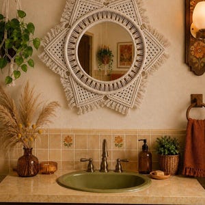 May include: A round wall mirror framed by an intricate off-white macrame border with a star-shaped design, featuring knotted patterns and fringed edges. The mirror is centered above a bathroom vanity with a green sink and a beige tiled backsplash.