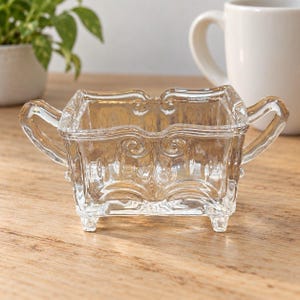 May include: A clear glass sugar bowl featuring a rectangular body with two side handles, four small feet, and embossed scrollwork patterns along the exterior walls. The vessel has an open top and a smooth, transparent finish throughout its structure.