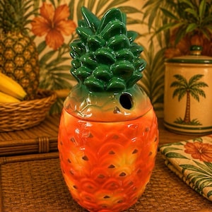 May include: A ceramic pineapple-shaped jar with a removable lid. The body of the jar is a vibrant orange with a textured, scale-like pattern. The lid is shaped like a green pineapple crown with a small circular opening on the side of the base.
