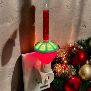 May include: A plug-in night light featuring a red bubble light tube atop a green and red plastic base. The device is inserted into a white wall outlet. The light emits a red glow, and the design includes a small toggle switch on the side.