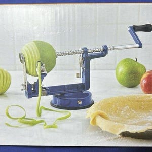 May include: A blue apple peeler with a black handle is peeling a green apple. A green apple is being peeled, with a long, green peel. There are two whole apples, one green and one red, and a pie crust in a pan.