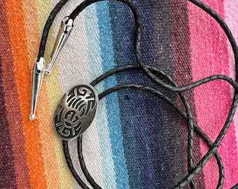 Op de afbeelding: Een zwarte leren bolo stropdas met zilverkleurige metalen punten en een decoratieve ovale hanger. De hanger heeft een uitgesneden ontwerp. De stropdas wordt gepresenteerd op een kleurrijk gestreept textiel.
