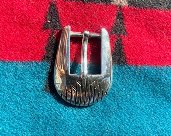 May include: A polished silver belt buckle featuring a rectangular bar and a decorative geometric pattern. The buckle is displayed on a colourful patterned textile background.