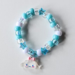 May include: A blue and white beaded bracelet with star-shaped charms and a white charm with a pink bow. The charm is a cartoon character with a smiling face.