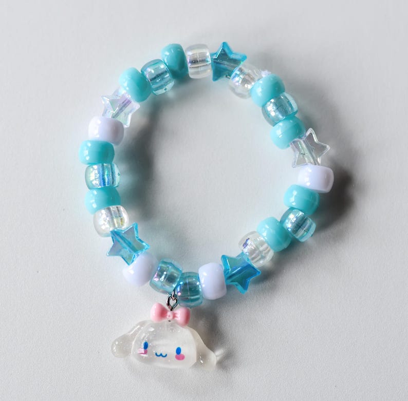 May include: A blue and white beaded bracelet with star-shaped charms and a white charm with a pink bow. The charm is a cartoon character with a smiling face.