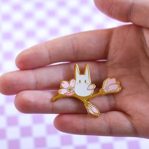 May include: A gold-colored enamel pin featuring a white cartoon bunny with long ears, perched on a branch with pink cherry blossoms. The pin is held in a hand against a soft purple and white checkered background.