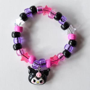 May include: A black and purple beaded bracelet with pink and clear star-shaped beads. A black and white charm of a cartoon character with a bow is attached to the bracelet.