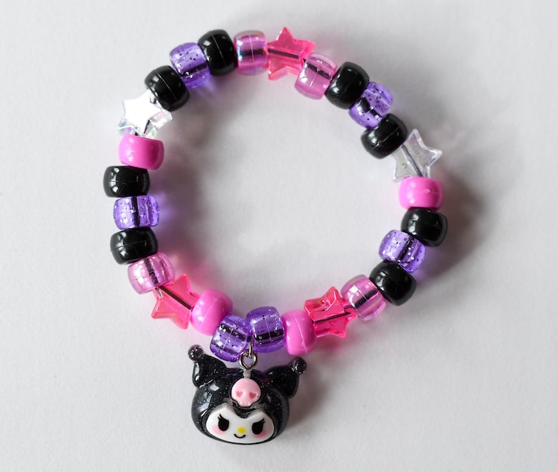 May include: A black and purple beaded bracelet with pink and clear star-shaped beads. A black and white charm of a cartoon character with a bow is attached to the bracelet.