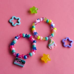 May include: Two beaded bracelets with star and charm accents, alongside star-shaped beads, on a pink background. The bracelets feature pink, blue, and yellow beads, with charms including a roller skate and a cassette tape.